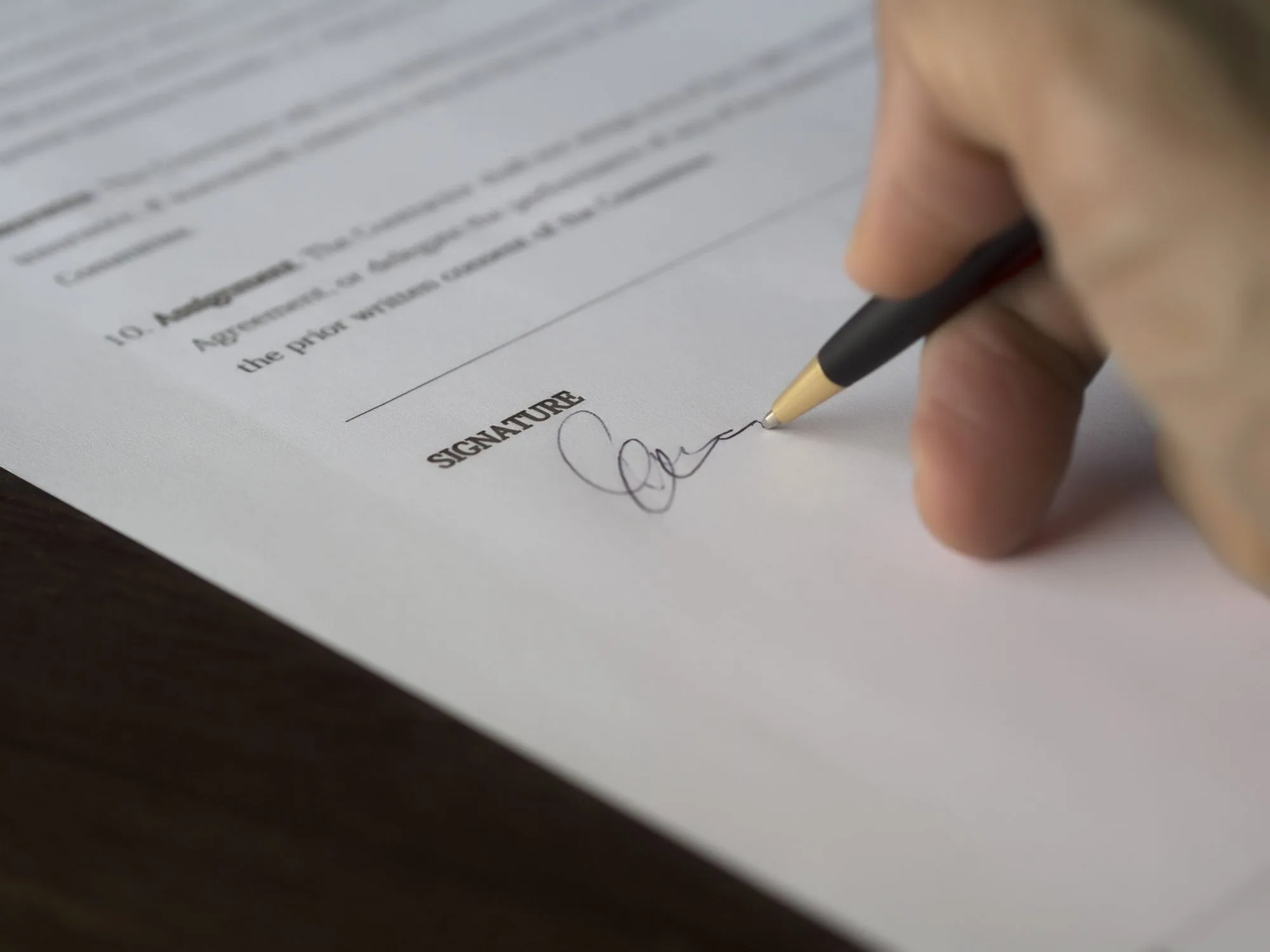 Una mano firmando un contrato con pluma sobre un escritorio de madera oscura