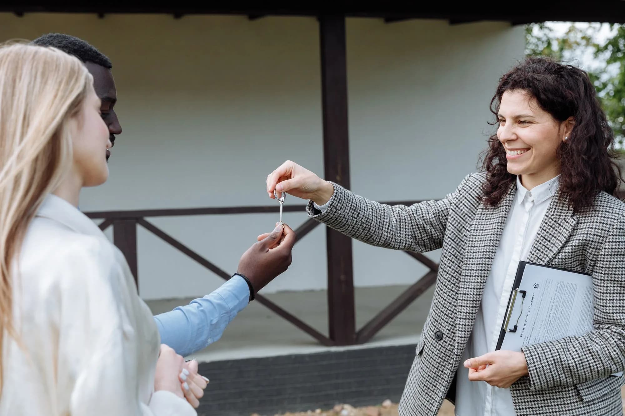 An agent handing a house key to a couple outside a white house with a timber porch