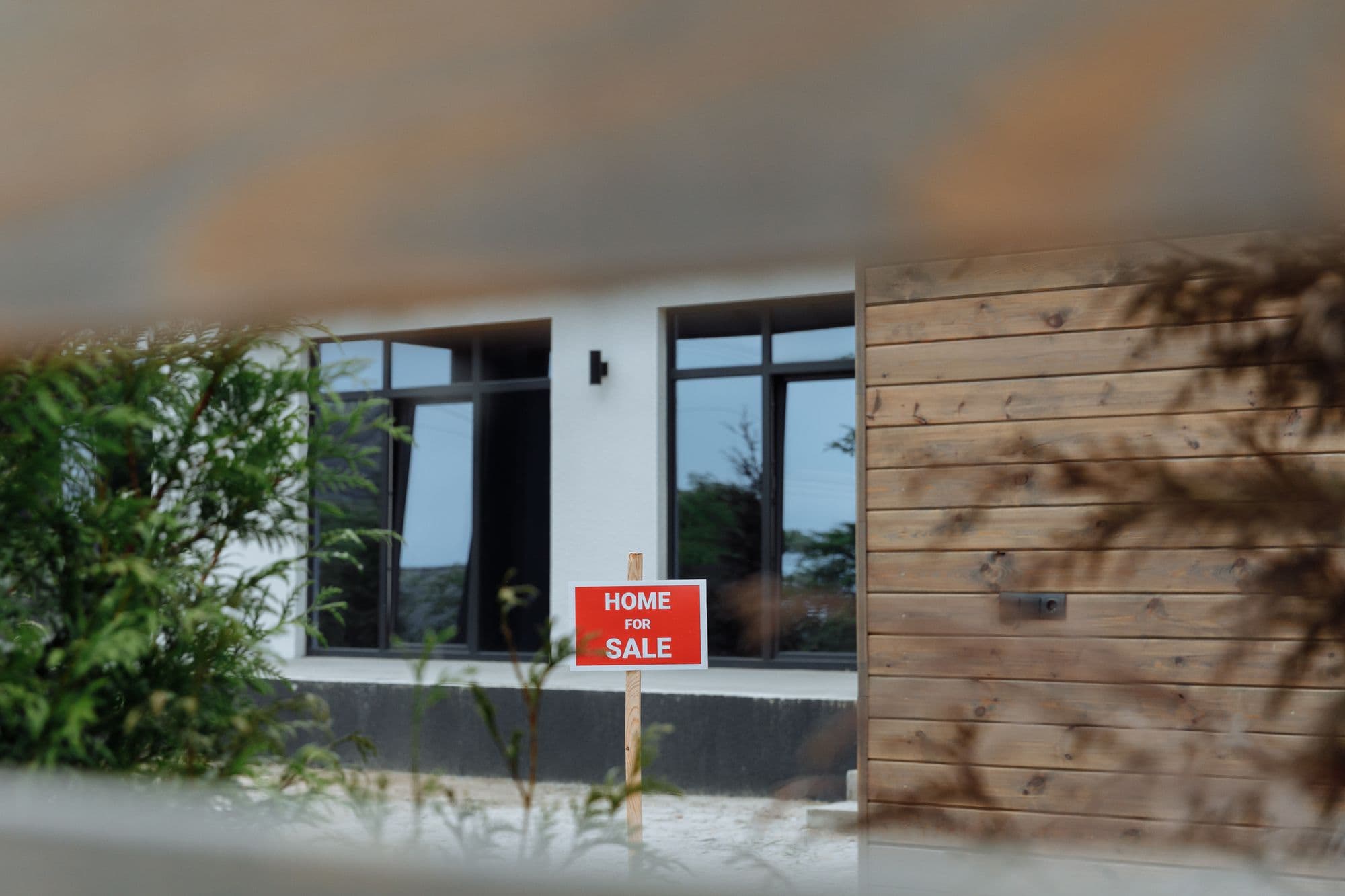 A contemporary house with a for-sale sign on the front lawn, framed by greenery