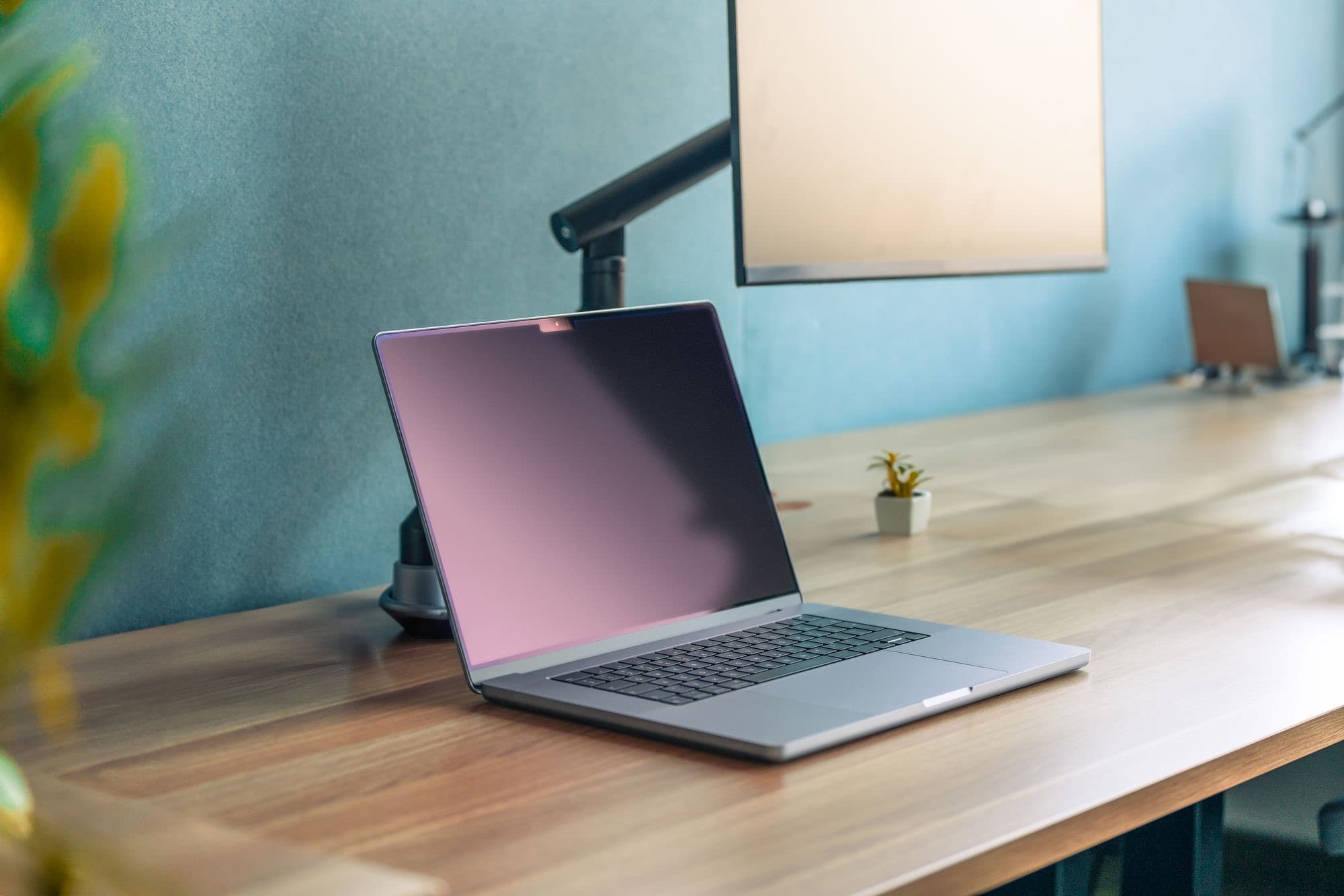 An open laptop on a wooden desk against a teal wall, a monitor on an arm above it