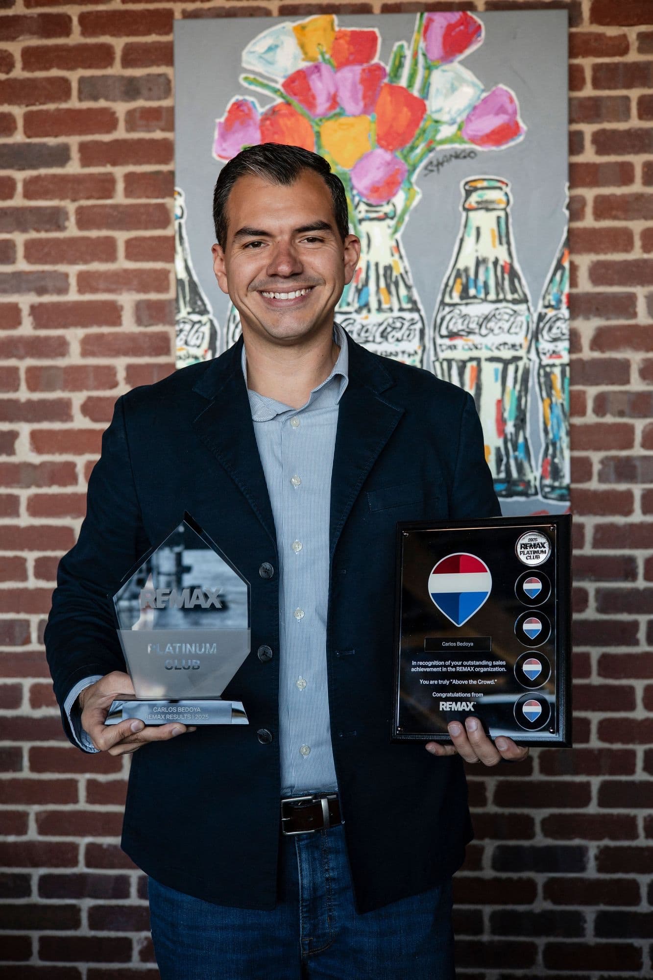 Carlos Bedoya sonriendo frente a una pared de ladrillo, con un premio y una placa del RE/MAX Platinum Club